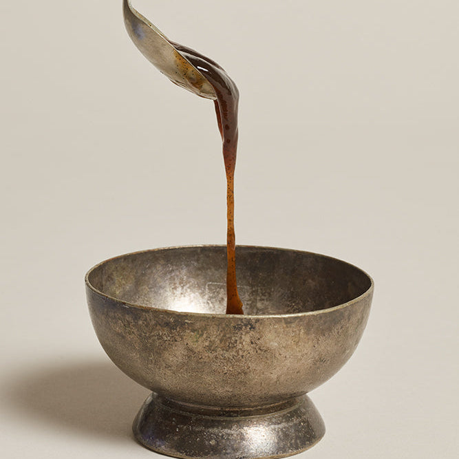 Vanilla bean paste being poured from a spoon into a metallic bowl on a neutral background