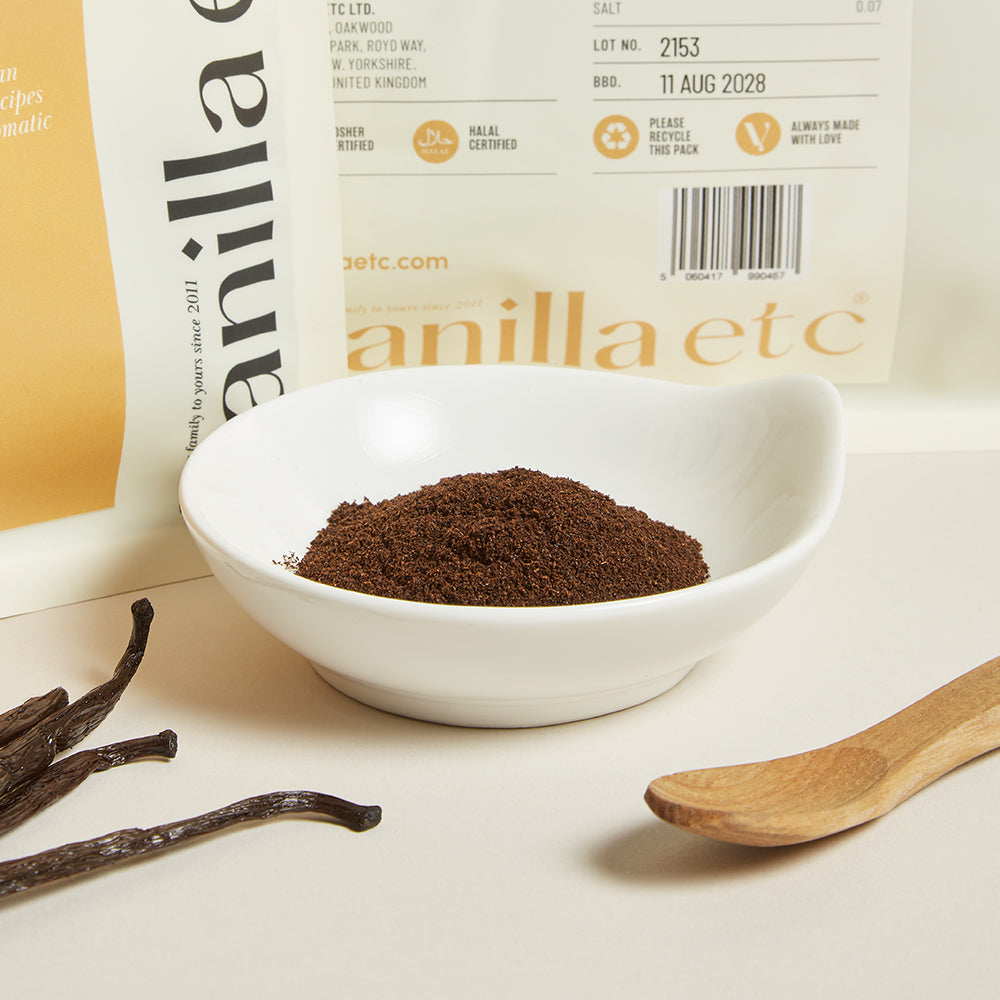 Vanilla powder in a white bowl with vanilla beans and a wooden spoon, against a vanilla etc branded background.
