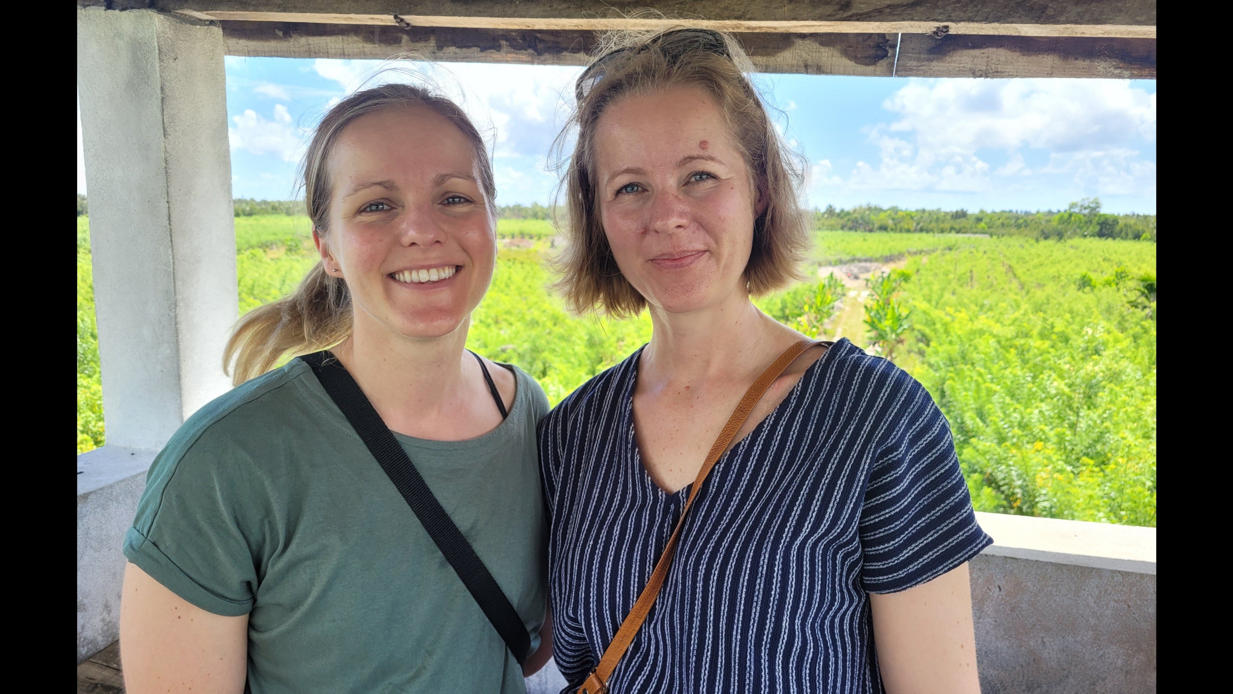 Two women standing together with a scenic view of a green vanilla plantation and blue sky in the background.