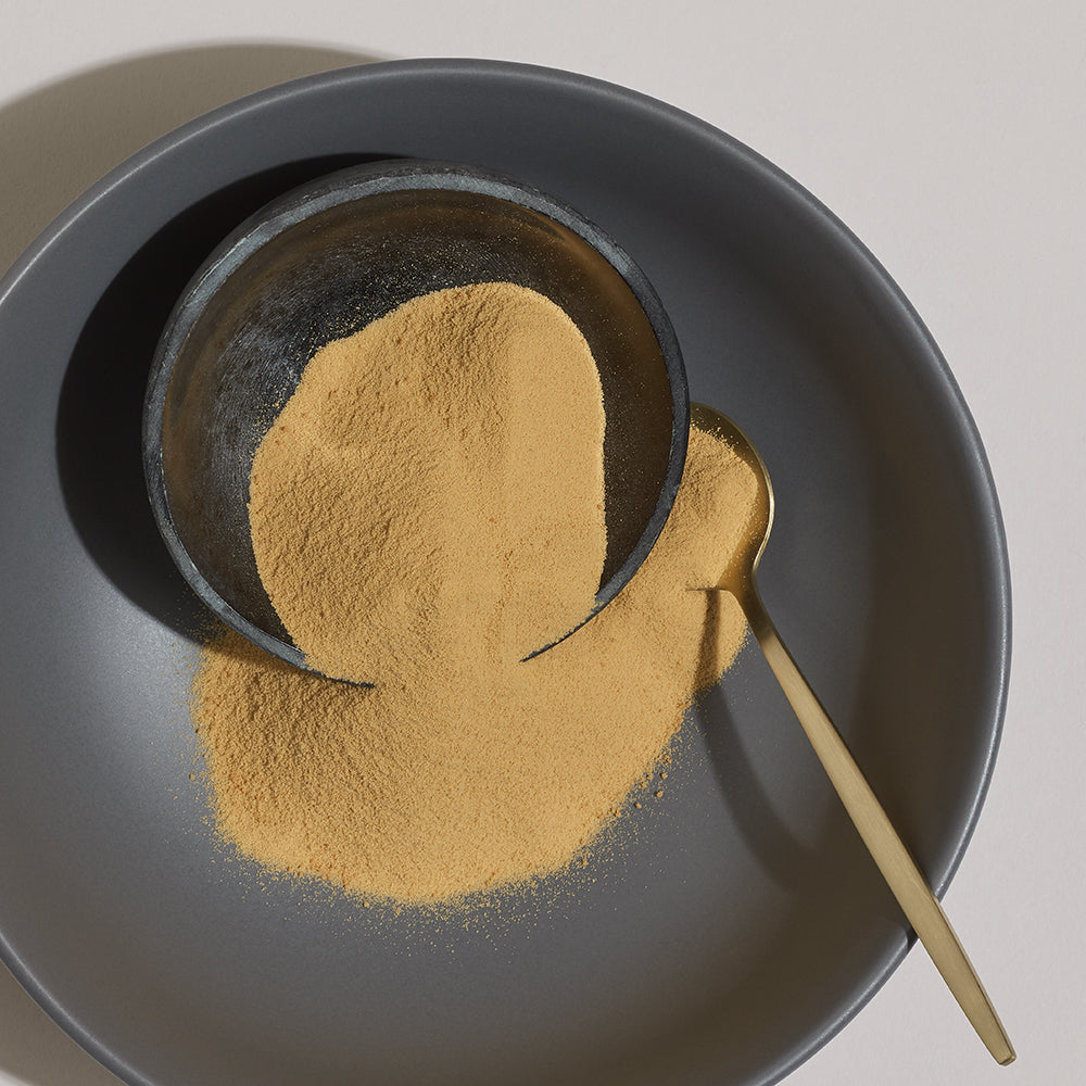 Golden powder in a black bowl on a grey plate with a wooden spoon.