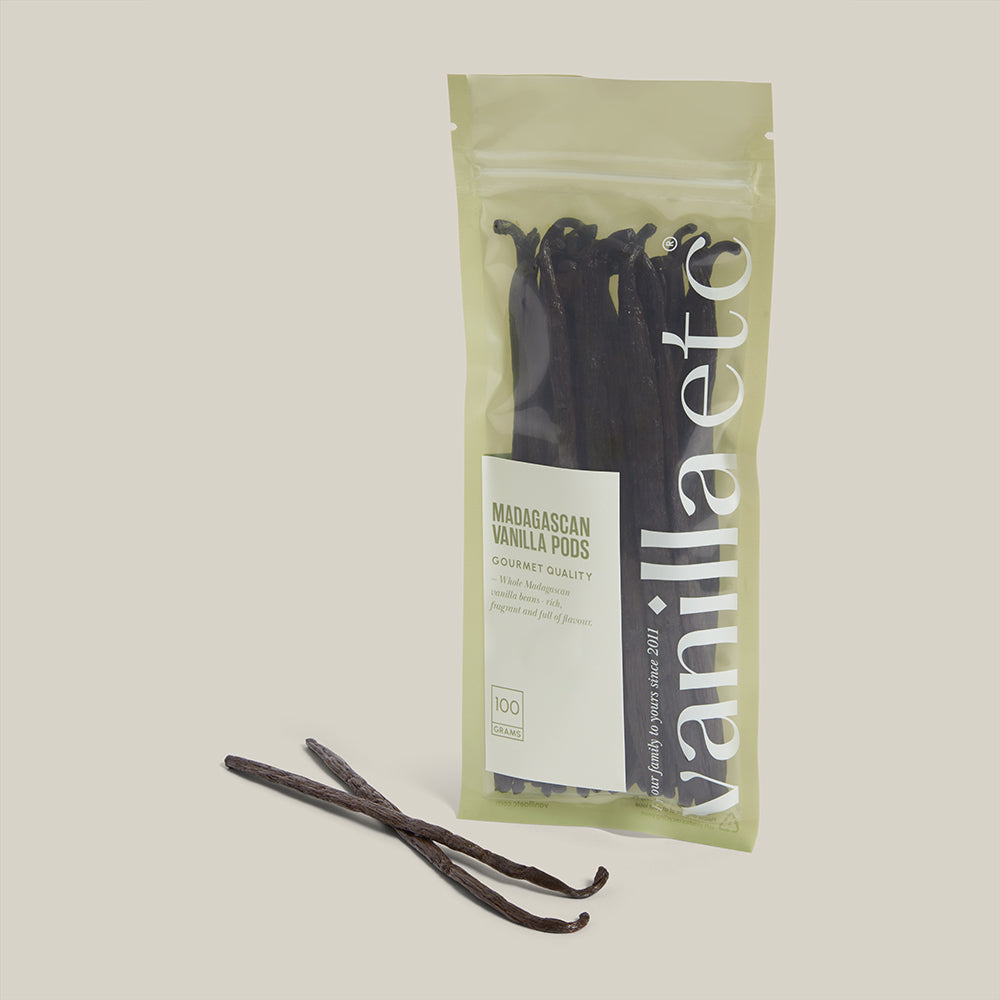 Packaged vanilla pods with a visible brand label on a neutral background and two beans without packaging in the foreground