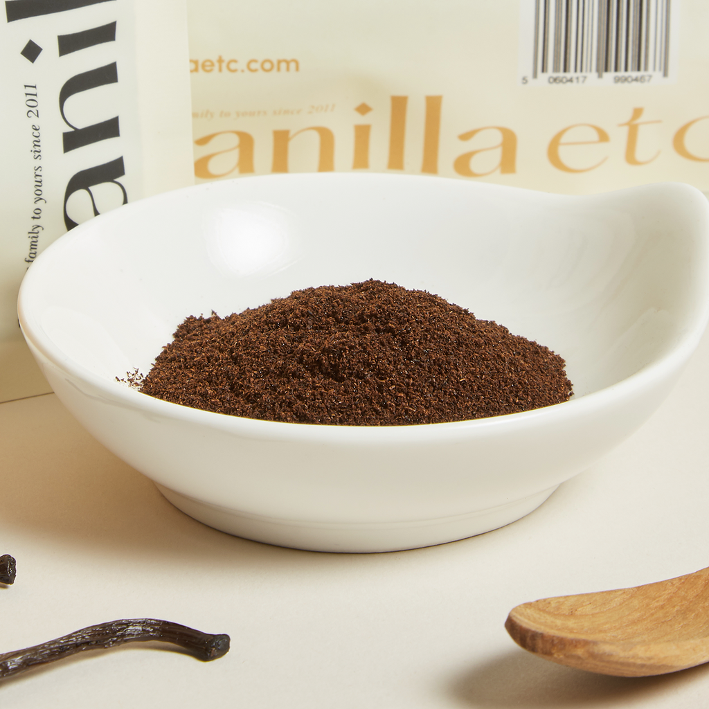 Vanilla powder in a white bowl with vanilla beans and a wooden spoon on a light background.