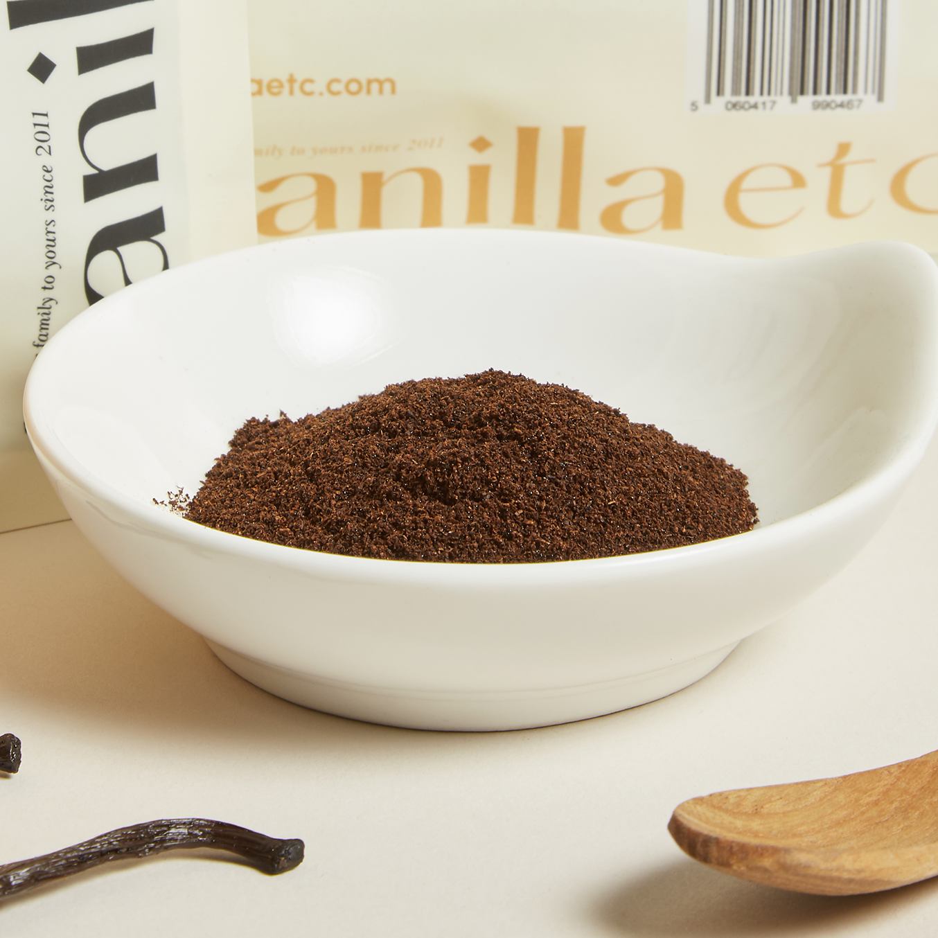 Vanilla powder in a white bowl with vanilla beans and a wooden spoon on a light background.