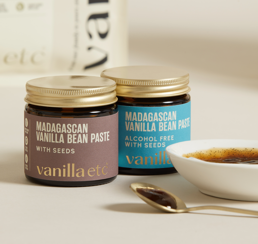 Two jars of Madagascan Vanilla Bean Paste with Seeds on a light surface, with a ceramic bowl and silver spoon showing consistency